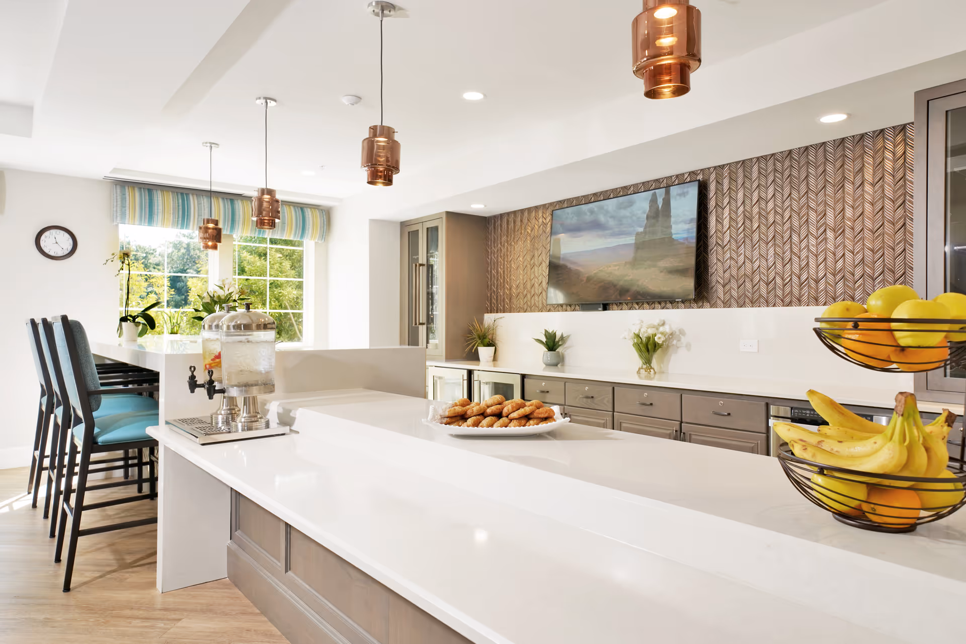 Bright and modern kitchen area with a long white countertop, bar stools with teal cushions, a water dispenser, a plate of croissants, and a two-tier fruit basket containing bananas and lemons. The background features a decorative wall with a mounted flat-screen TV, cabinets, and potted plants. Large windows with striped valances allow natural light to fill the space.