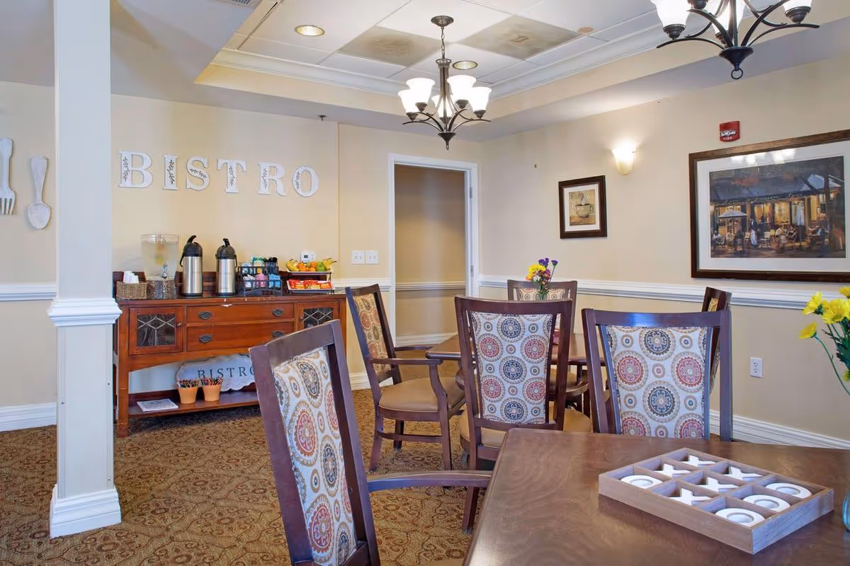 A cozy dining area in a senior living facility with wooden tables and chairs featuring patterned cushions. A wooden sideboard against the wall holds coffee dispensers, a water container, and snacks. The wall behind the sideboard has the word 'BISTRO' displayed in decorative letters. The room is softly lit with ceiling lights and wall sconces, and framed artwork hangs on the walls. A tic-tac-toe game is placed on one of the tables.