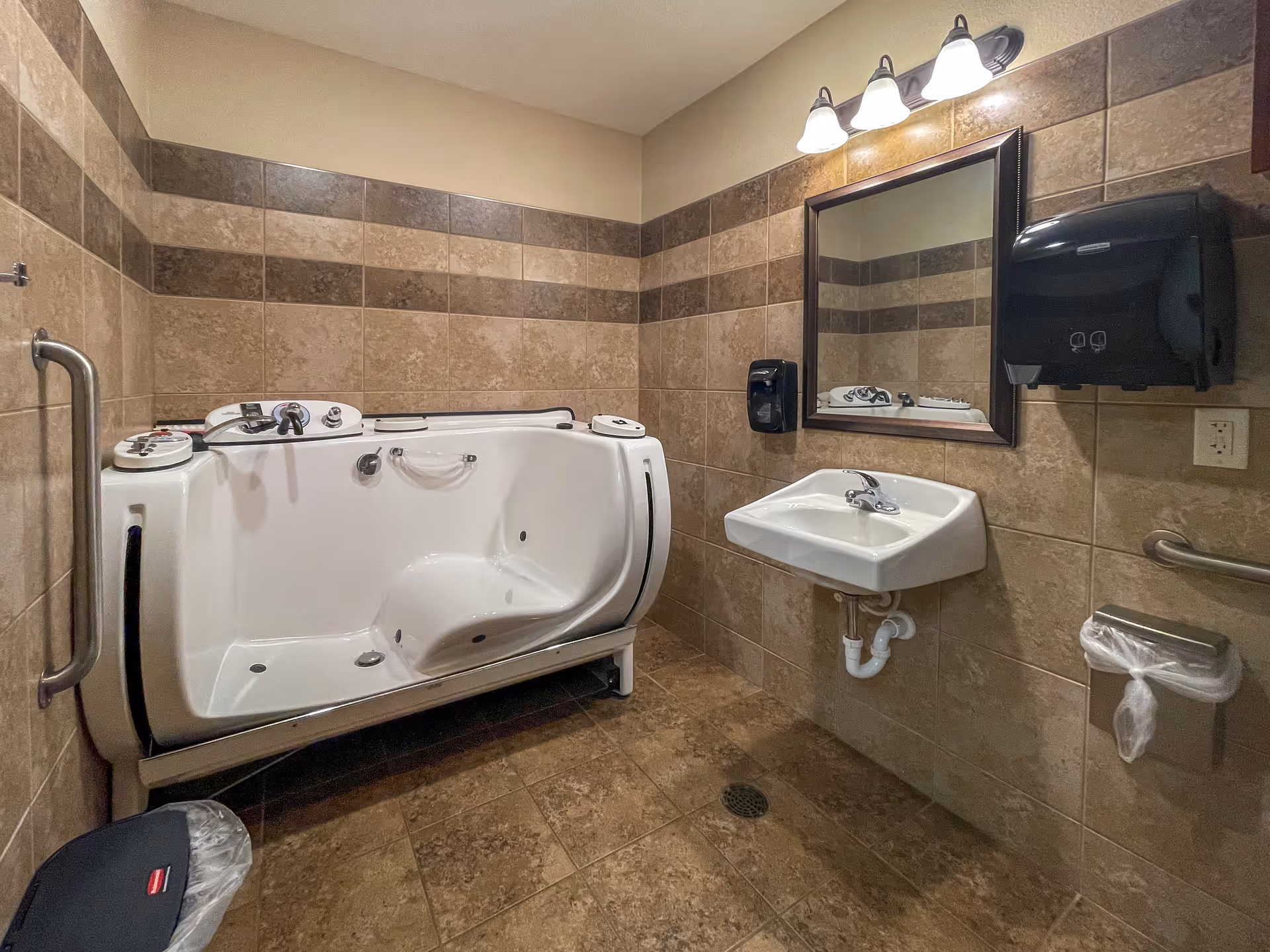 A bathroom with beige and brown tiled walls and floor, featuring a walk-in bathtub with safety handles, a wall-mounted sink with a mirror above it, a soap dispenser, a paper towel dispenser, a trash can, and grab bars for accessibility.