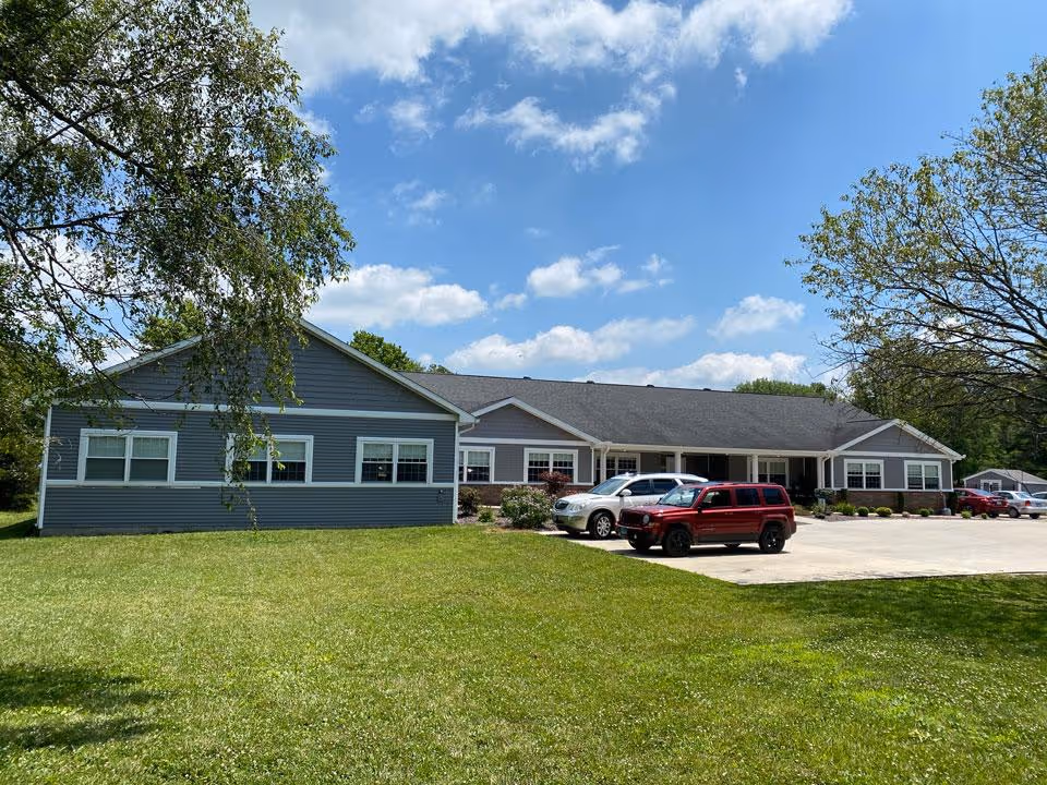 Exterior view of a single-story gray building with white trim, surrounded by green grass and trees under a blue sky with scattered clouds. Several cars are parked in front of the building.