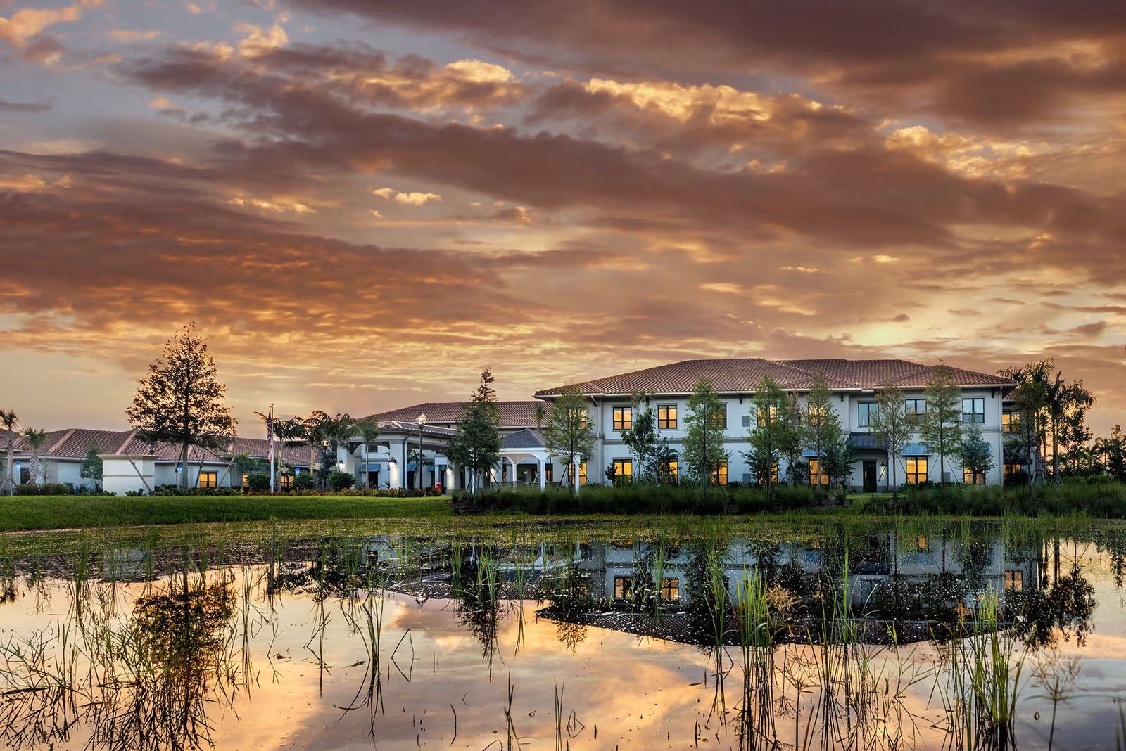 A two-story senior living building at sunset with warm interior lights, reflected in a pond in the foreground.