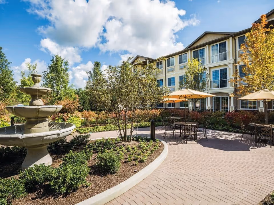 Outdoor patio area at StoryPoint Pickerington featuring a multi-tiered stone fountain, landscaped garden beds with shrubs and small trees, paved walkways, and several tables with umbrellas for shade. The background shows a multi-story residential building under a partly cloudy blue sky.