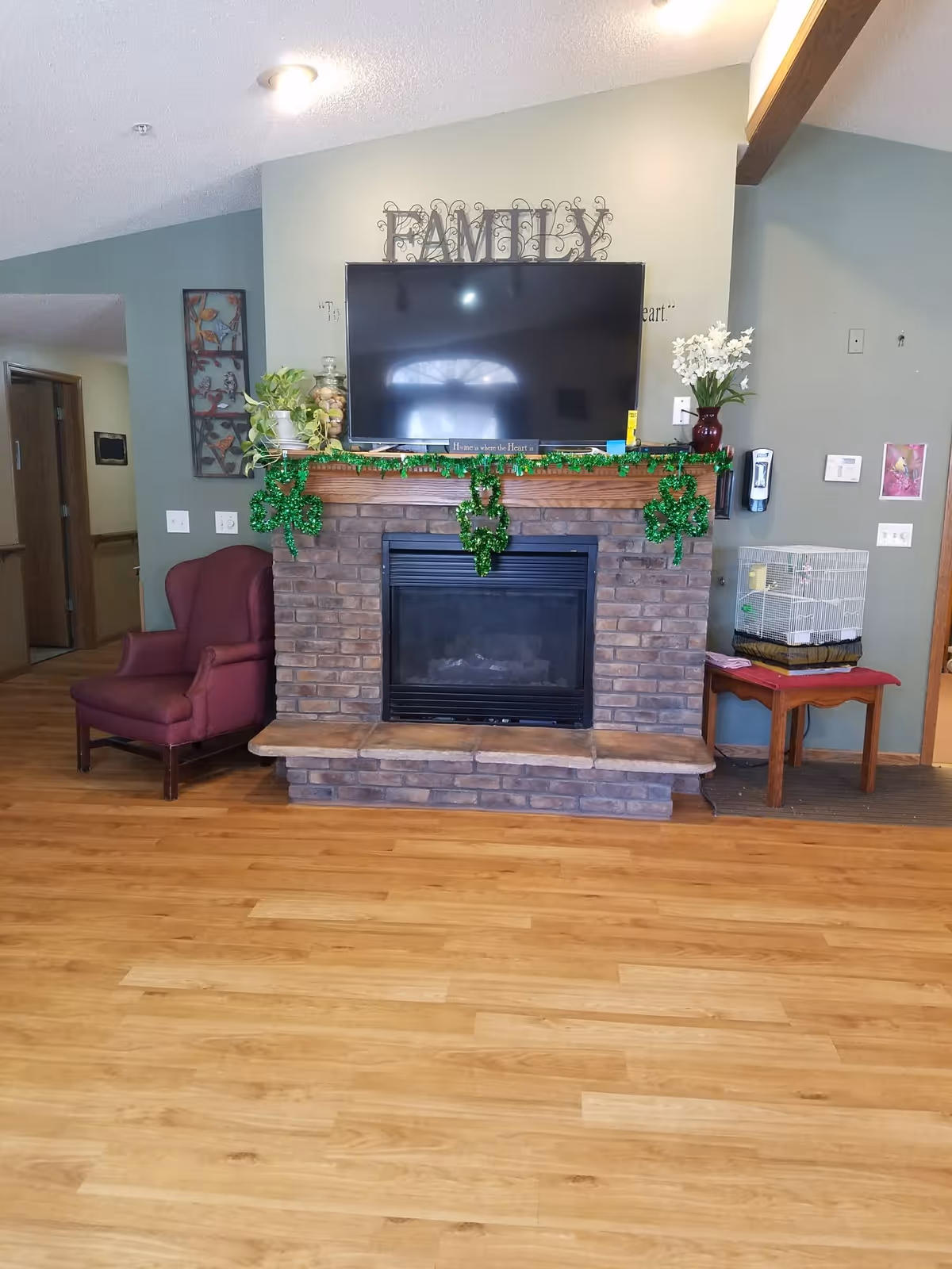 A cozy living room area featuring a brick fireplace with a mounted flat-screen TV above it. The mantel is decorated with green shamrock garlands and plants. To the left of the fireplace is a maroon armchair, and to the right is a small wooden table with a birdcage on top. The walls are painted in muted green and beige tones, and the floor is light wood.