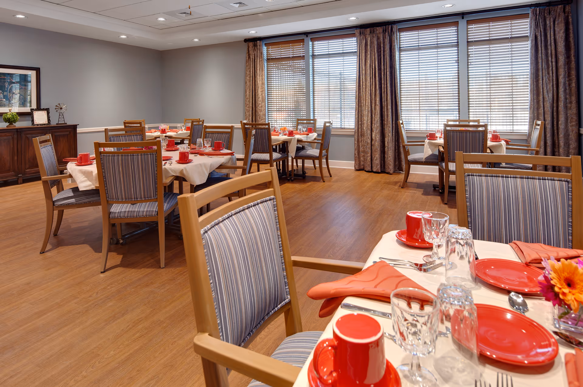 A dining room with several tables set for a meal. Each table has white tablecloths, red plates, red cups, orange napkins, and glassware. The chairs have wooden frames with striped upholstery. Large windows with blinds and patterned curtains allow natural light into the room. A wooden sideboard with decorative items is visible against one wall.