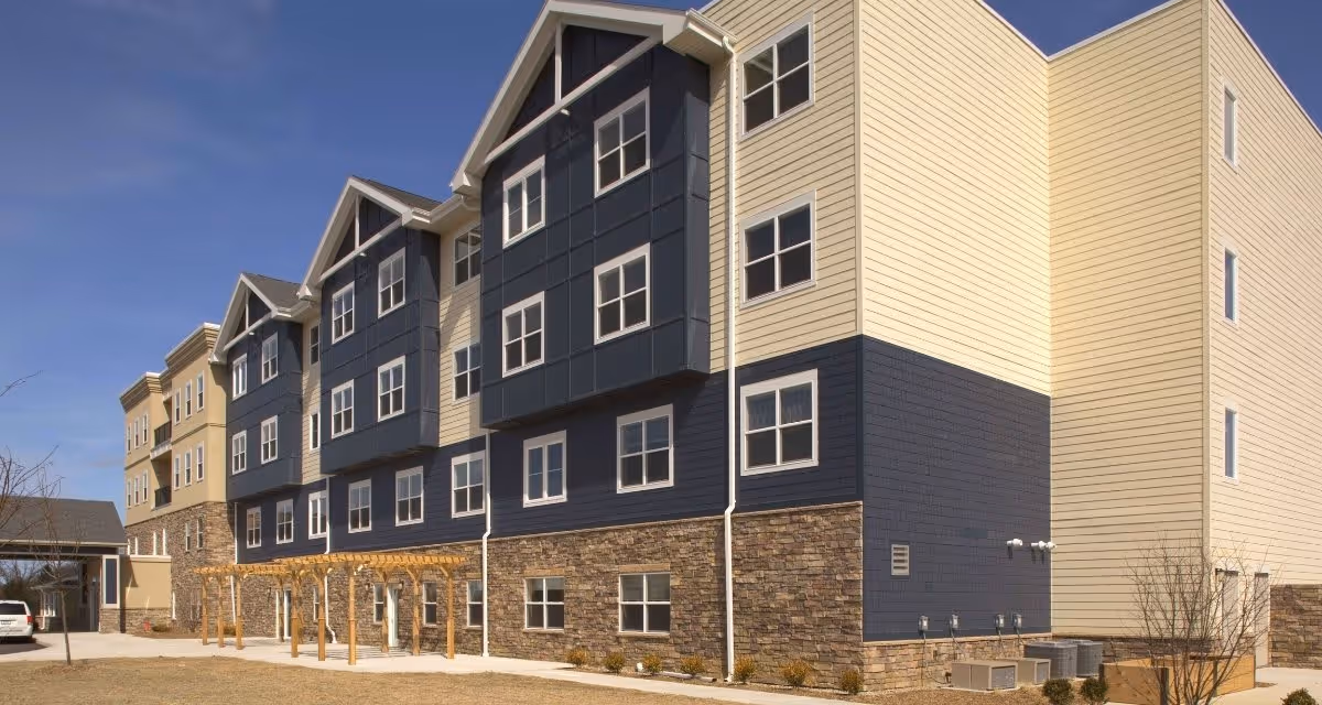 Exterior view of a multi-story senior living facility building with beige, navy blue, and stone facade under a clear blue sky. There is a covered entrance on the left side and a wooden pergola structure along the sidewalk.
