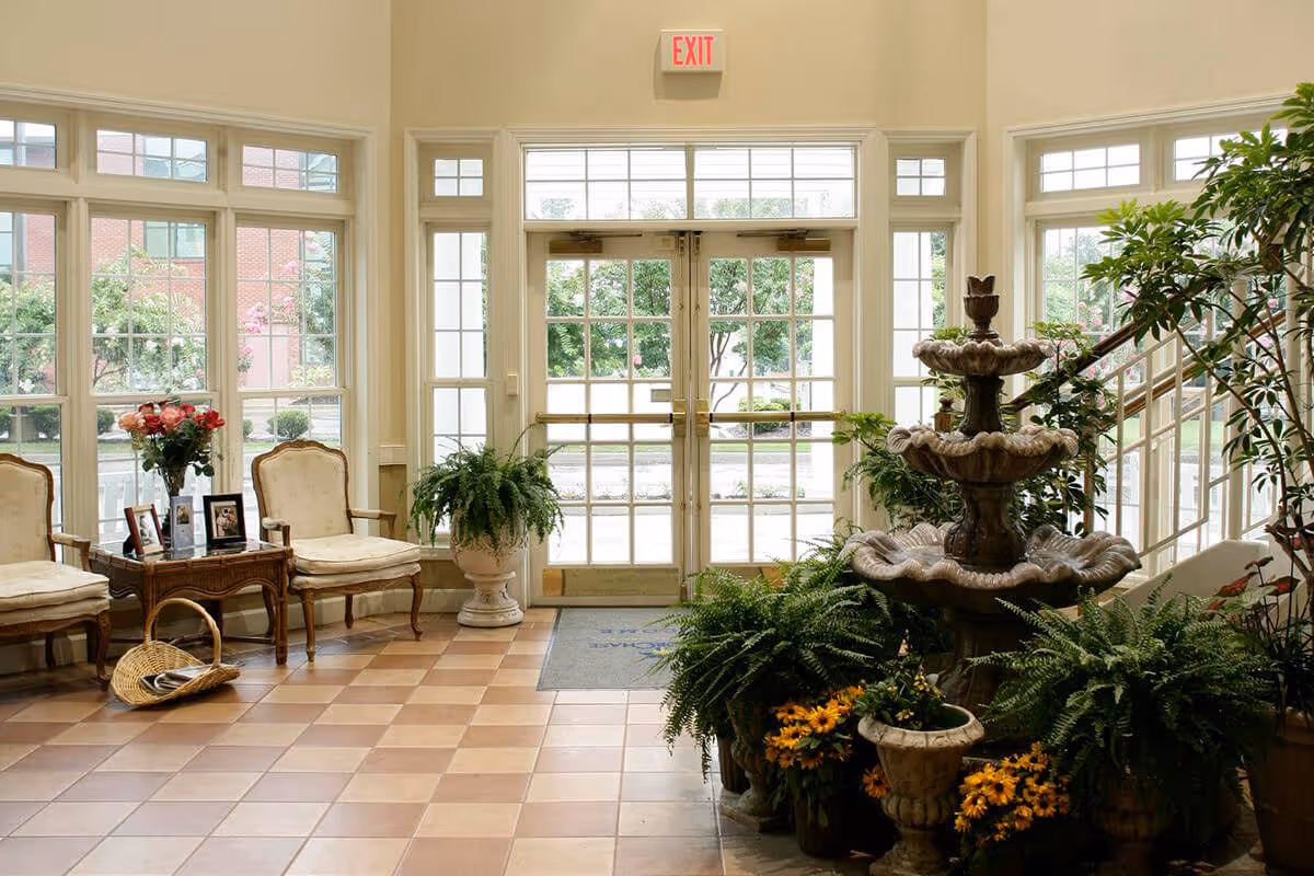 Bright lobby entrance with glass double doors, a tiered fountain surrounded by plants, and chairs beside large windows.