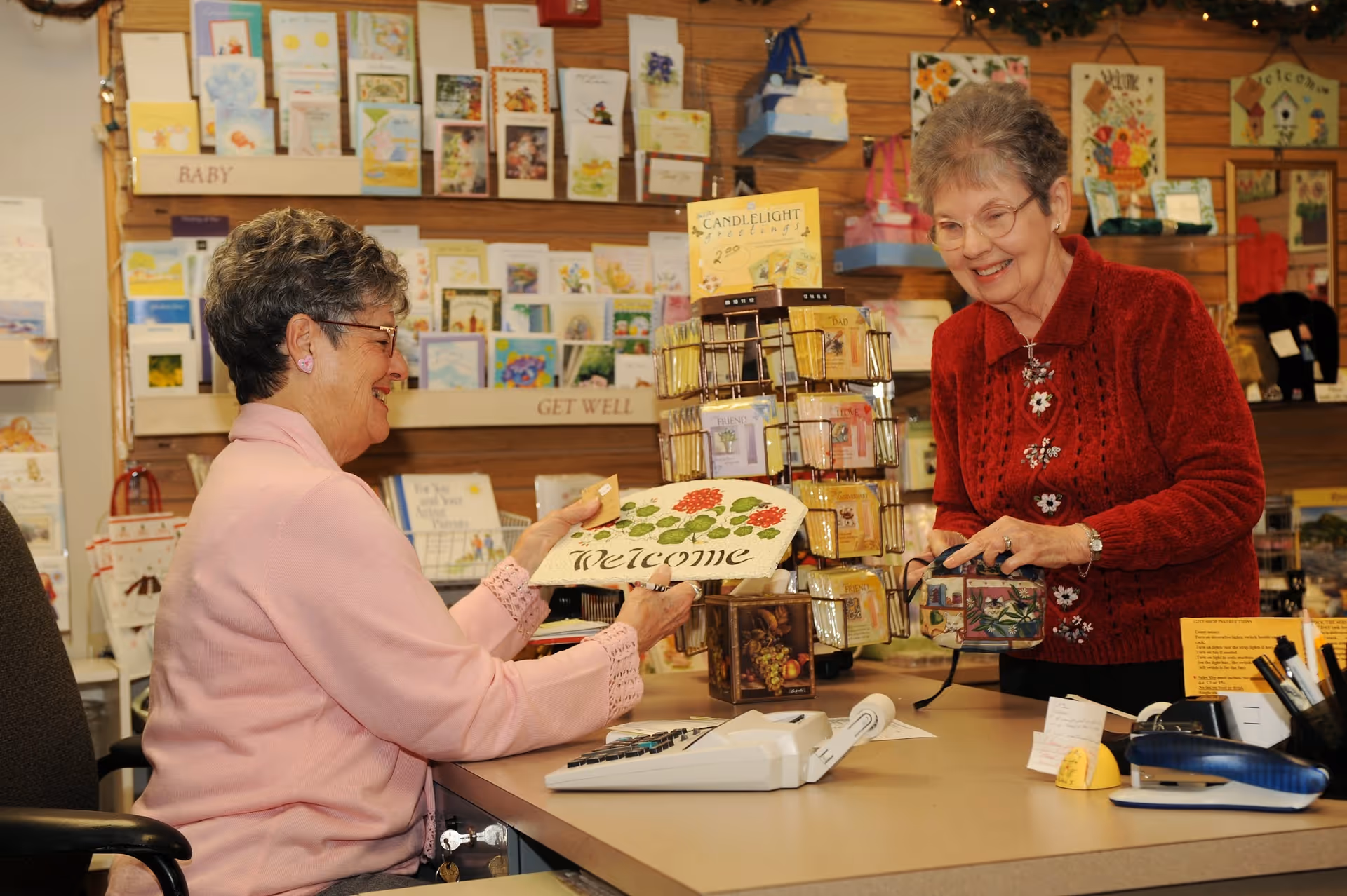 Two elderly women smiling and interacting at a counter inside a gift shop area. One woman in a pink sweater is holding a decorative 'Welcome' sign, while the other woman in a red sweater is holding a small purse. Behind them are shelves filled with greeting cards and small gift items.