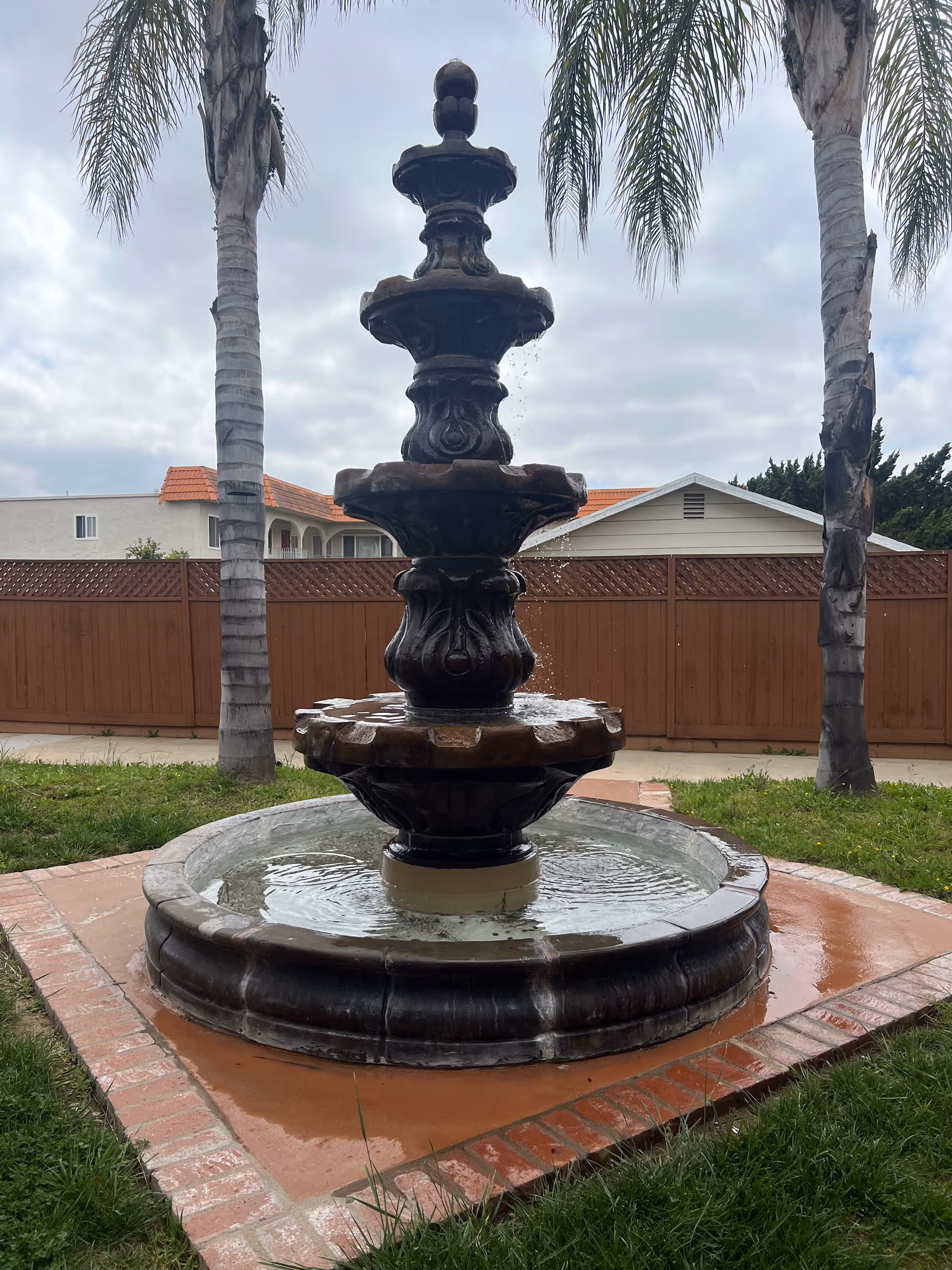 A multi-tiered decorative water fountain made of dark stone, situated outdoors on a brick and concrete base surrounded by grass. Two tall palm trees flank the fountain, and a wooden fence with houses in the background is visible under a cloudy sky.