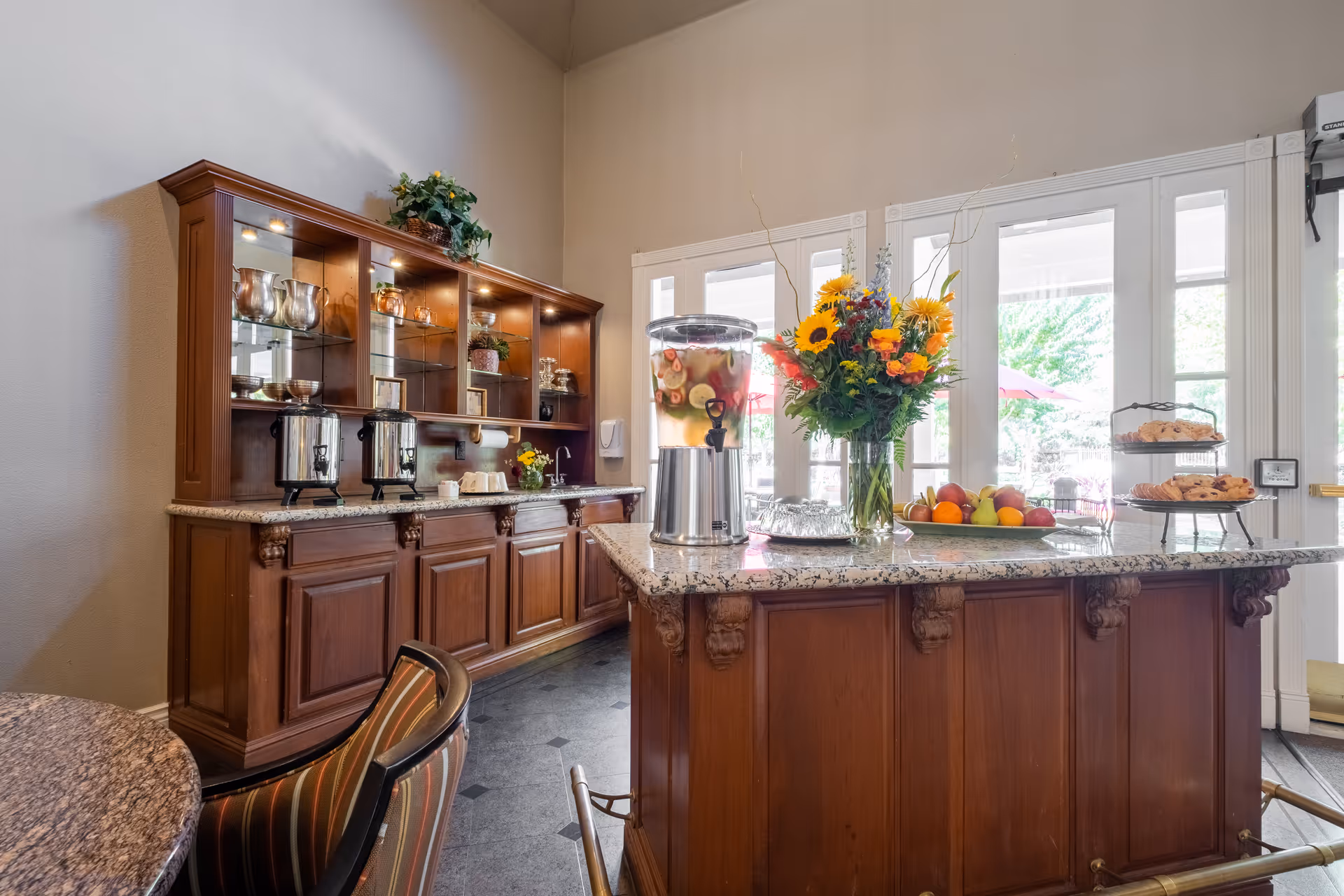 Interior view of a dining or common area in a senior living facility with a wooden cabinet and countertop. The countertop holds a large beverage dispenser filled with fruit-infused water, a vase with a colorful flower arrangement, a plate of fresh fruit, and a tiered tray with pastries. Behind the counter, there are glass shelves with decorative items and two coffee urns. Large windows and glass doors let in natural light and show an outdoor patio with umbrellas.