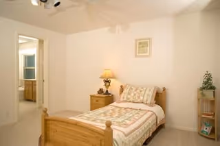 A small bedroom with a single wooden bed covered with a patterned quilt and pillow. Next to the bed is a wooden nightstand with a lamp and decorative items. There is a framed picture hanging on the wall above the bed. To the left, an open door leads to a bathroom with a visible vanity and mirror. A small wooden shelf with plants and books is on the right side of the room.