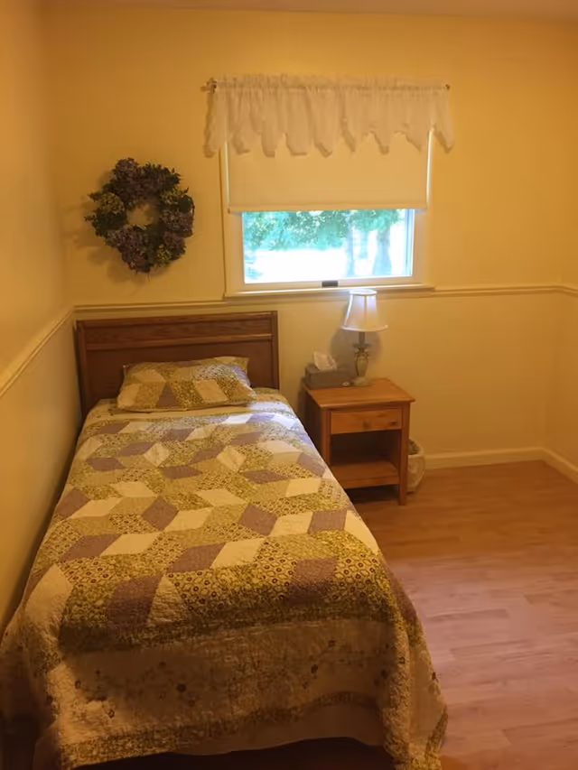 A small bedroom with a single bed covered with a patchwork quilt in shades of green, purple, and beige. There is a wooden headboard and a matching wooden nightstand beside the bed with a lamp and a box of tissues on it. A window with a white roller shade and lace valance is above the nightstand, and a decorative wreath hangs on the wall to the left of the window. The room has light-colored walls and wood flooring.
