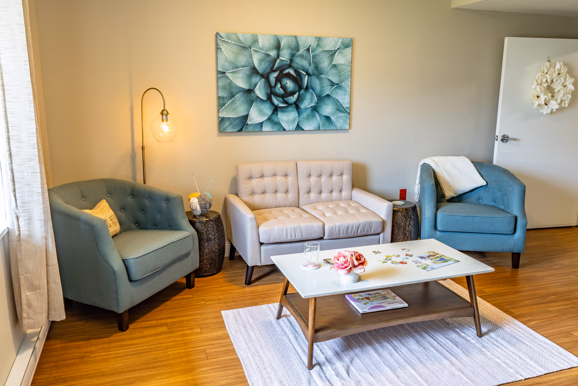 Cozy living room with two teal armchairs, a beige loveseat, a coffee table, and wall art above the sofa.