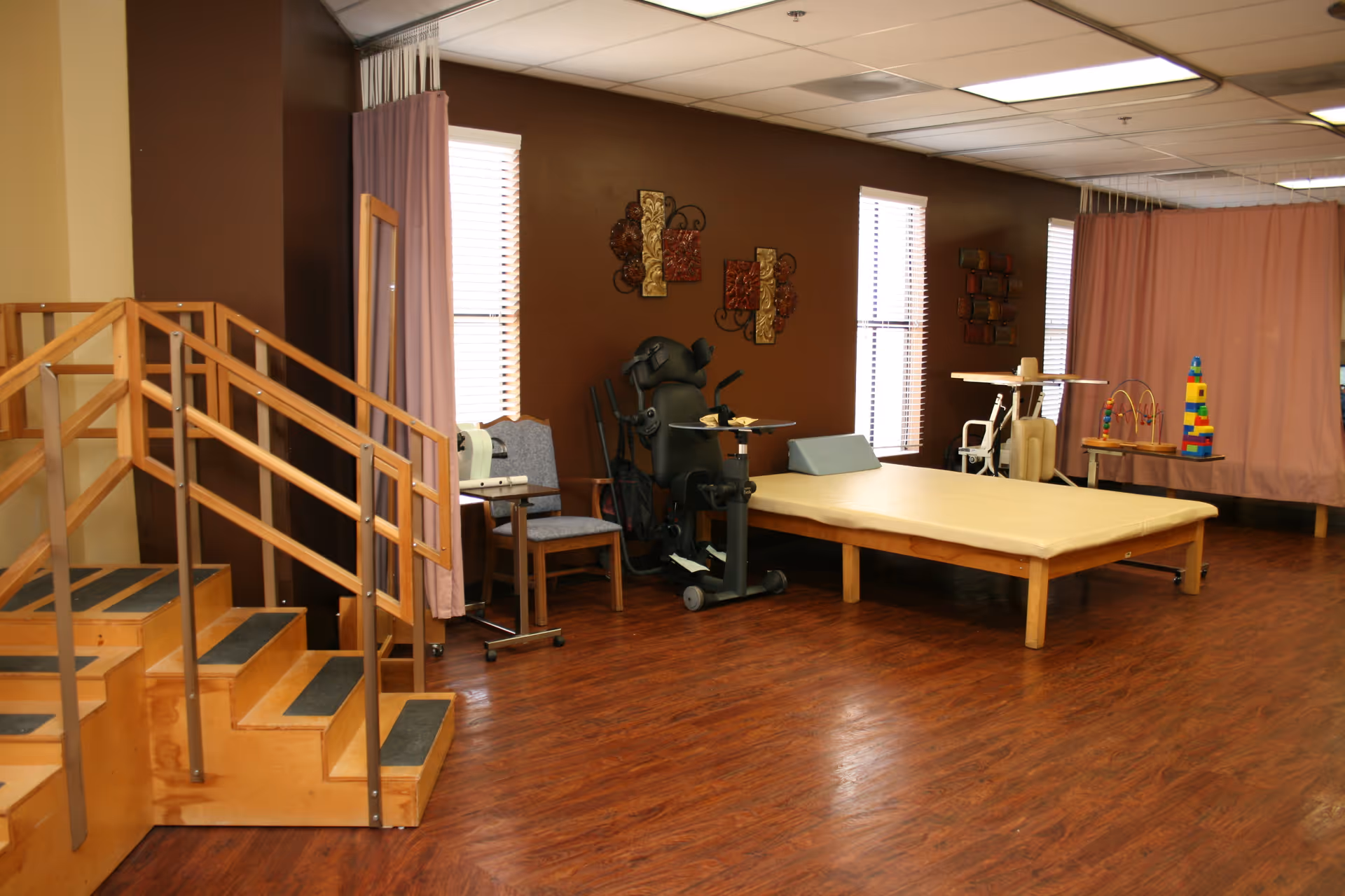 A therapy or rehabilitation room with wooden stairs equipped with handrails, a padded therapy table, exercise equipment, chairs, and therapy toys. The room has brown walls, wood flooring, and windows with blinds and curtains.