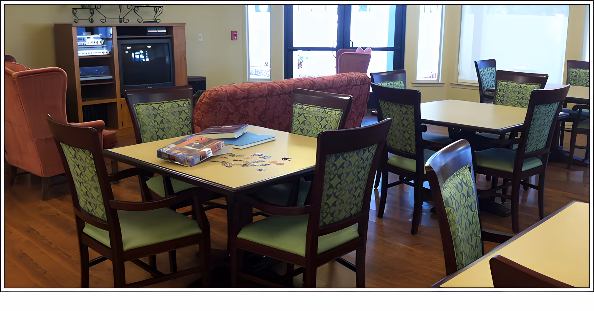 A common area in a senior living facility with several tables and chairs arranged for group activities. One table has a partially completed jigsaw puzzle and some books on it. The room has wooden flooring, large windows letting in natural light, and comfortable upholstered chairs and sofas in the background.