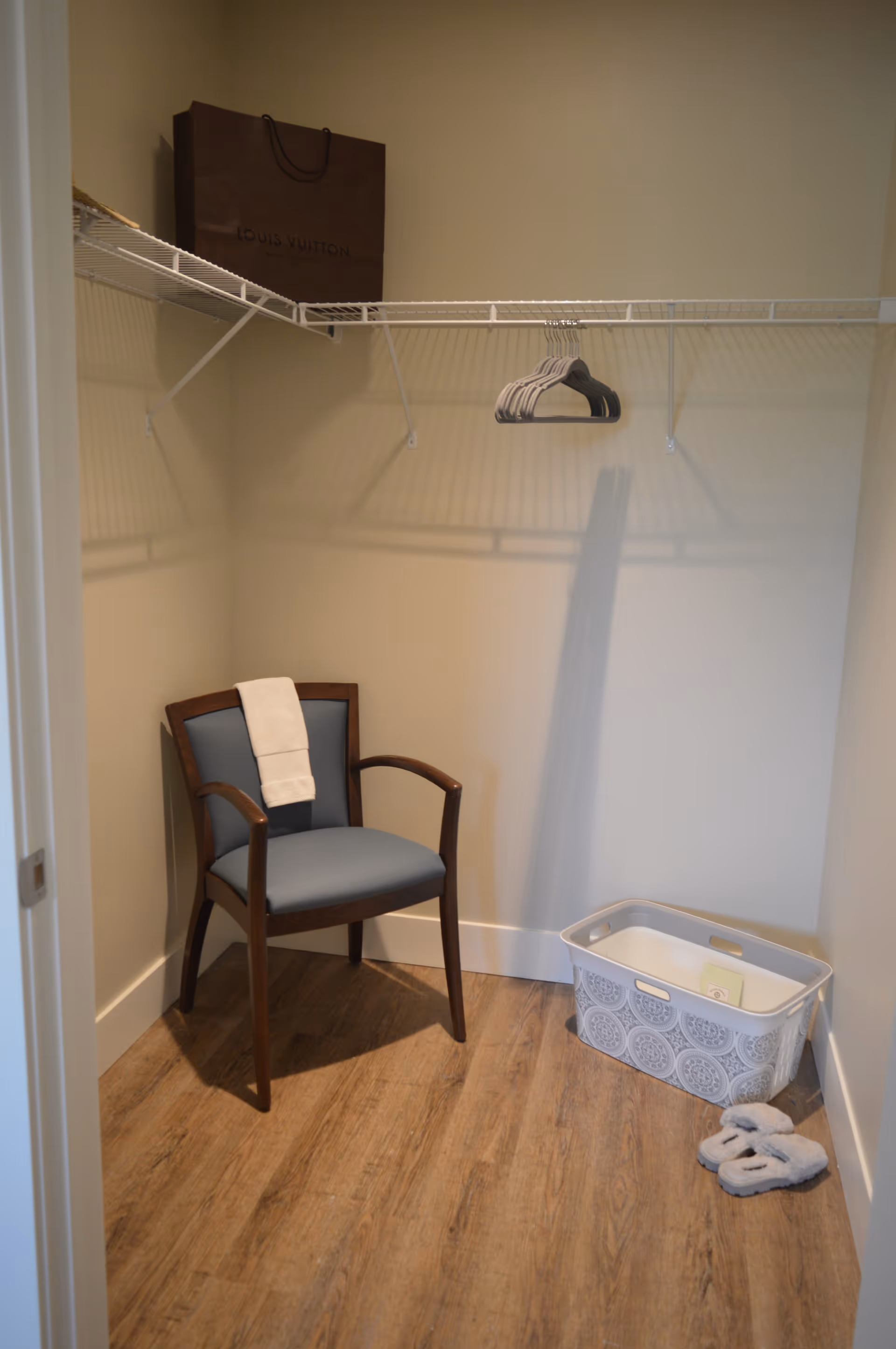 A small walk-in closet with wooden flooring, a wooden chair with a blue cushion and a white towel draped over the back, a white laundry basket with a decorative pattern, a pair of gray slippers on the floor, and a brown Louis Vuitton shopping bag on a white wire shelf with several empty hangers.