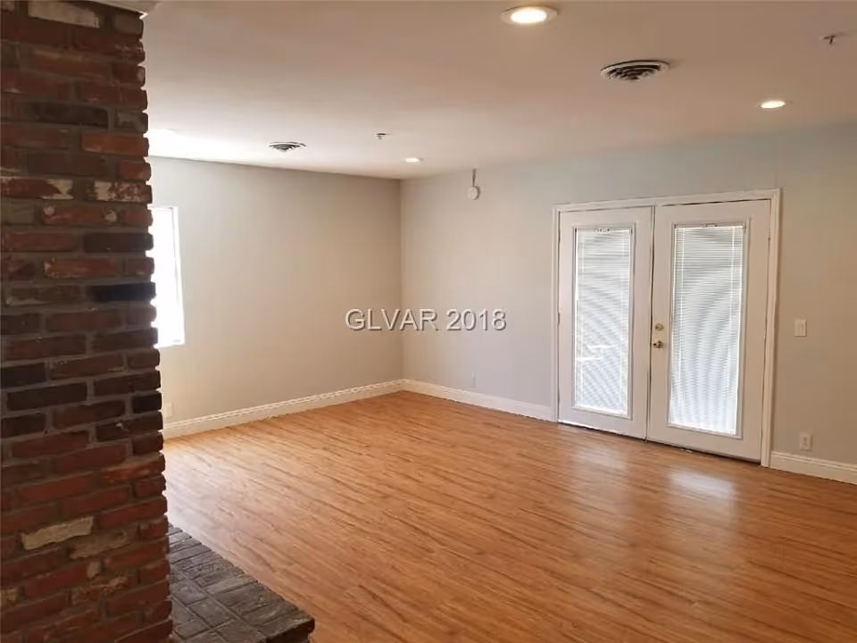Empty room with light wood flooring, white walls, a brick fireplace on the left, a window on the left wall, and double glass doors with blinds on the right wall.