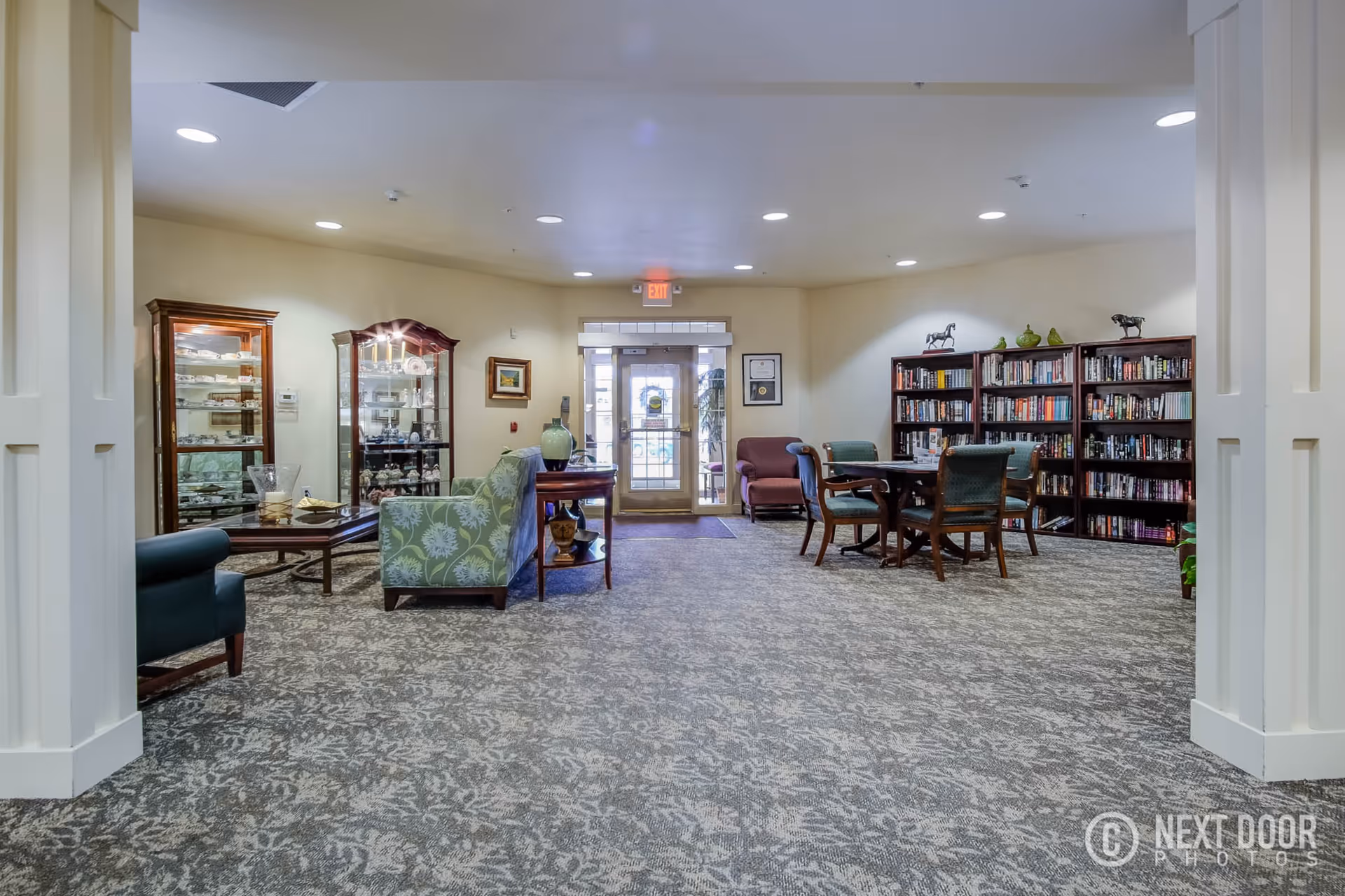 A spacious and well-lit common area in a retirement village featuring patterned carpet flooring, comfortable upholstered chairs and sofas, wooden display cabinets with decorative items, a table with chairs, and bookshelves filled with books. The area has a glass door exit with an exit sign above it.
