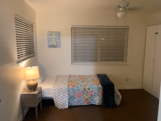 A simple bedroom with a single bed covered in a floral quilt, a nightstand with a lamp, a window with blinds, ceiling fan, and a wardrobe.