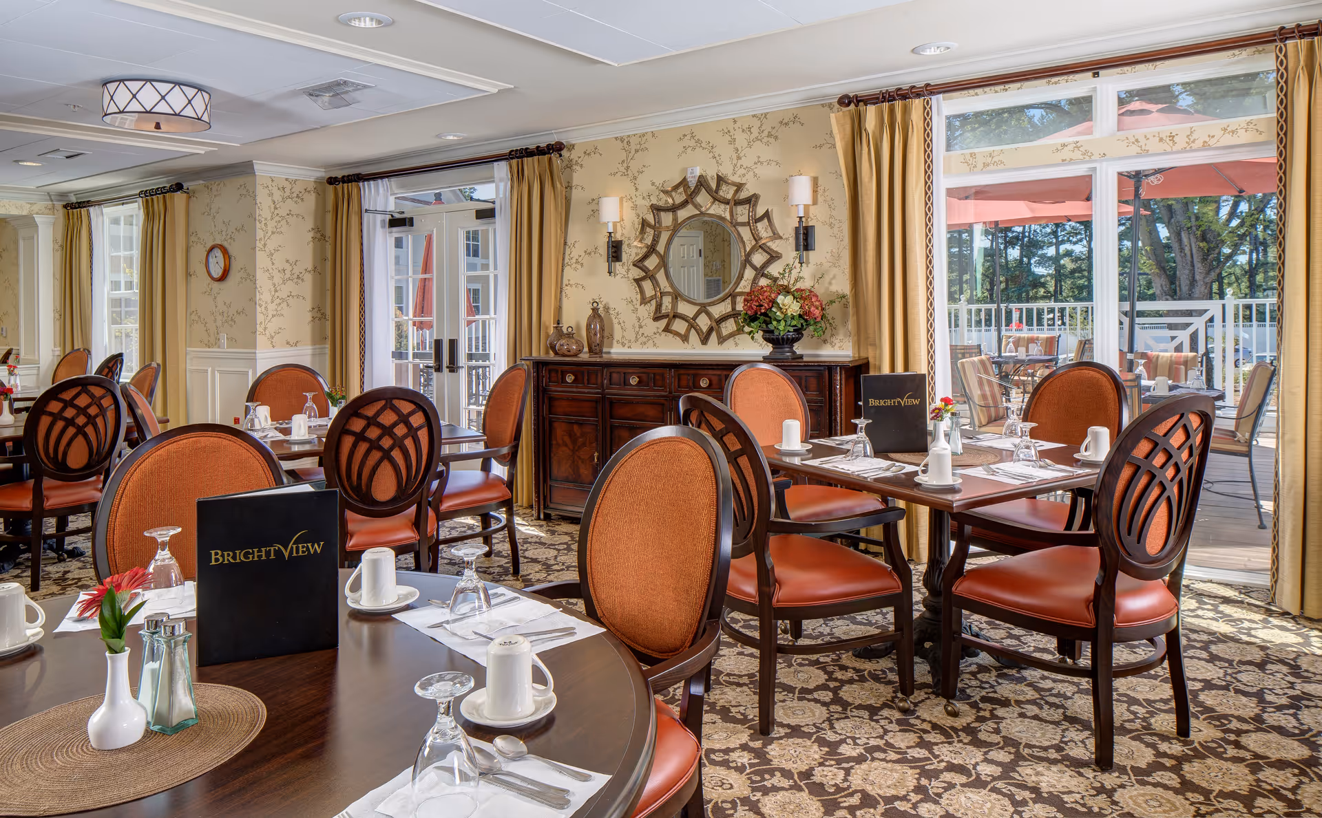 A bright and elegant dining room with round and square wooden tables set with white cups, glasses, and silverware. The chairs have orange cushioned seats and backs with dark wooden frames. There are menus labeled 'Brightview' on some tables. The room has patterned carpet, wallpaper with a subtle floral design, and large windows with gold curtains that open to an outdoor patio with additional seating and red umbrellas.