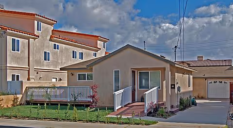 Exterior view of a single-story residential building with a small front porch and ramp, adjacent to a multi-story building under a partly cloudy sky.