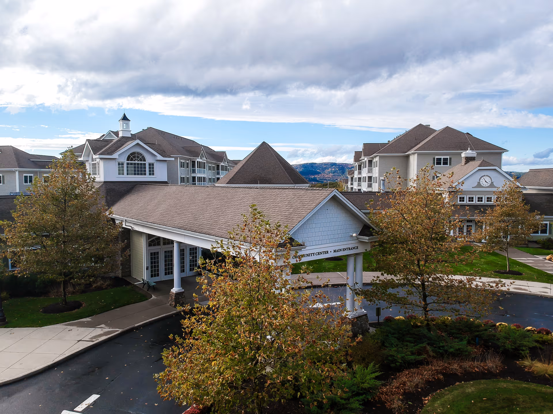 Exterior view of Woodland Pond At New Paltz senior living facility showing the community center main entrance with a covered driveway, surrounded by trees with autumn foliage and multi-story residential buildings in the background under a partly cloudy sky.
