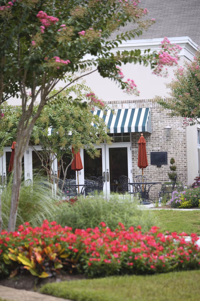 Outdoor patio area with black metal tables and chairs, red umbrellas, and a striped green and white awning above glass doors. The patio is surrounded by lush greenery, trees, and vibrant pink flowers in the foreground.