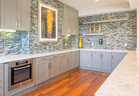 Modern kitchen with gray cabinets, marble countertops, mosaic tile backsplash, and wood flooring.