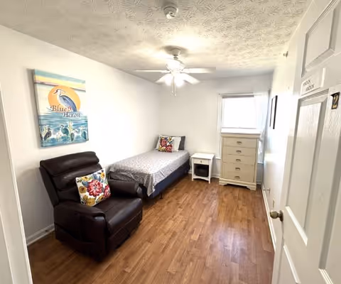 A small, simple bedroom with a single bed against the far wall, a white nightstand next to it, a tall white dresser near a window with sheer curtains, and a dark brown recliner chair with a colorful floral pillow. A ceiling fan with a light is on the ceiling, and a painting of a blue heron hangs on the left wall. The floor is wooden, and the door to the room is partially open.