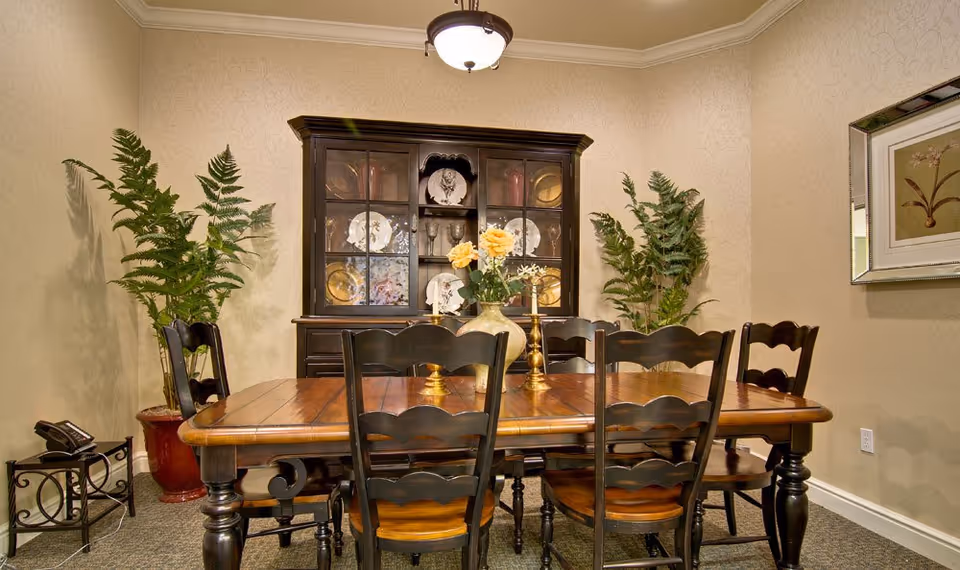 A formal dining room with a wooden table and chairs, a china cabinet, potted plants, and a vase of flowers on the table.