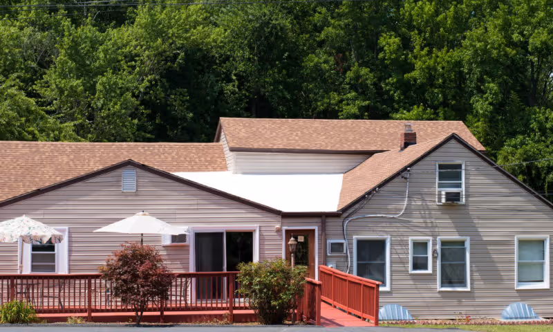 Exterior view of a single-story residential building with beige siding and a brown shingled roof, featuring a red wooden ramp and railing leading to the front door. There are two umbrellas providing shade on the porch area, surrounded by green bushes and trees in the background.