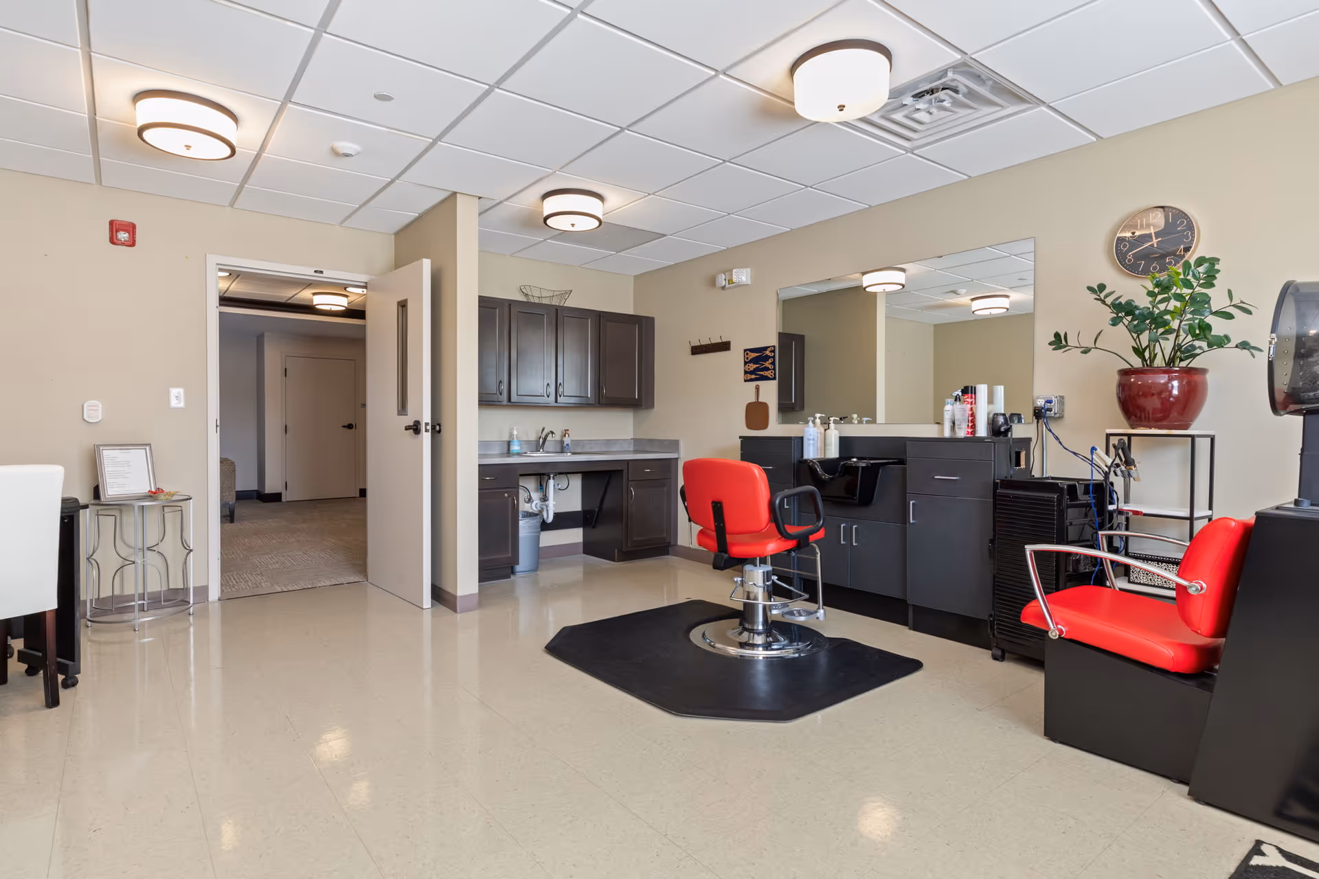 Bright salon-style room with red styling chairs, a sink and cabinets, a large mirror and potted plant.
