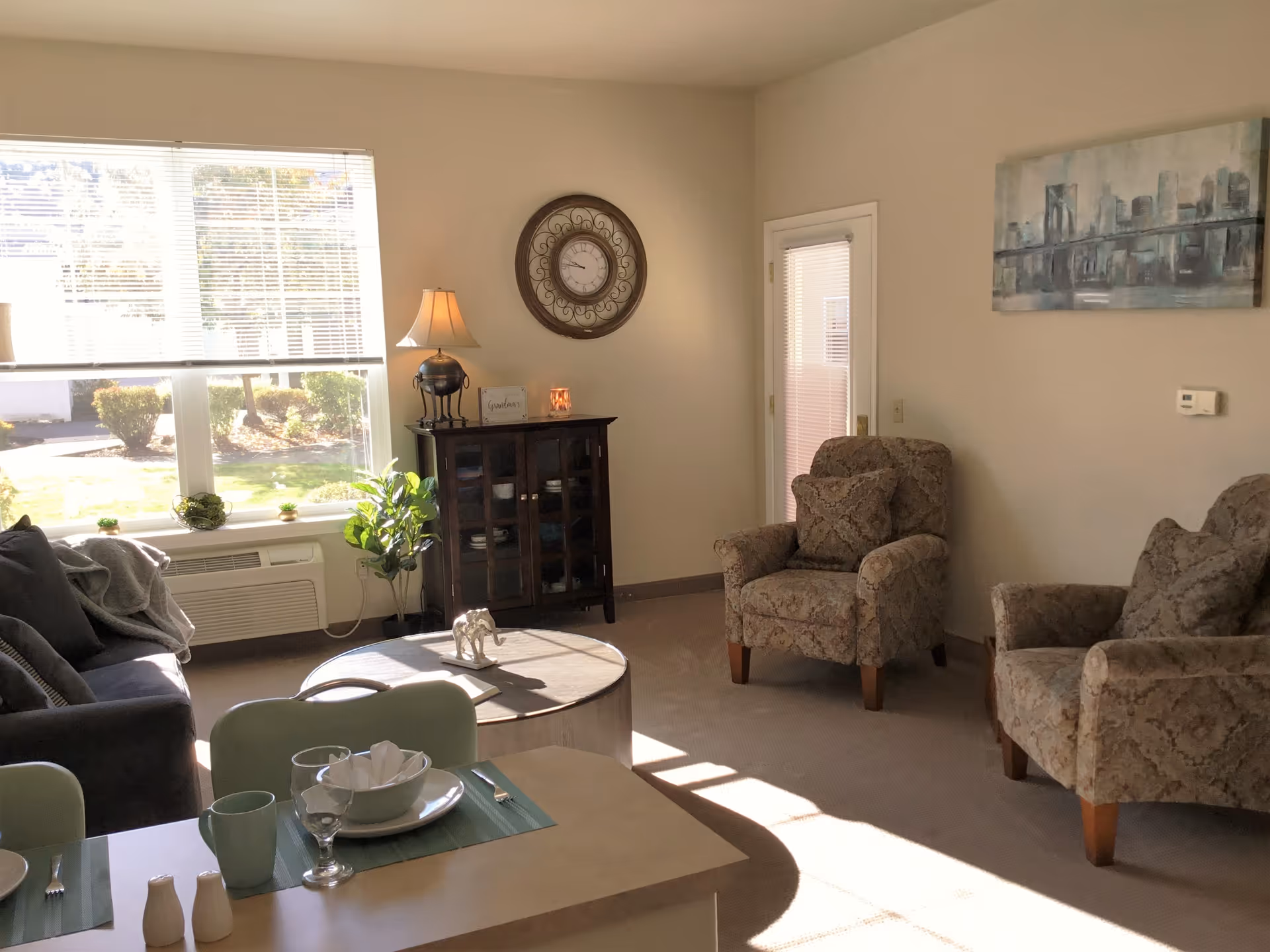 A sunlit living room with a large window covered by blinds, a dark sofa with cushions and a throw blanket, two patterned armchairs, a round wooden coffee table with a decorative elephant figurine, a dark wooden cabinet with a lamp and small decor items on top, a wall clock, a painting of a city skyline, and a dining table set with plates, bowls, glasses, and utensils.