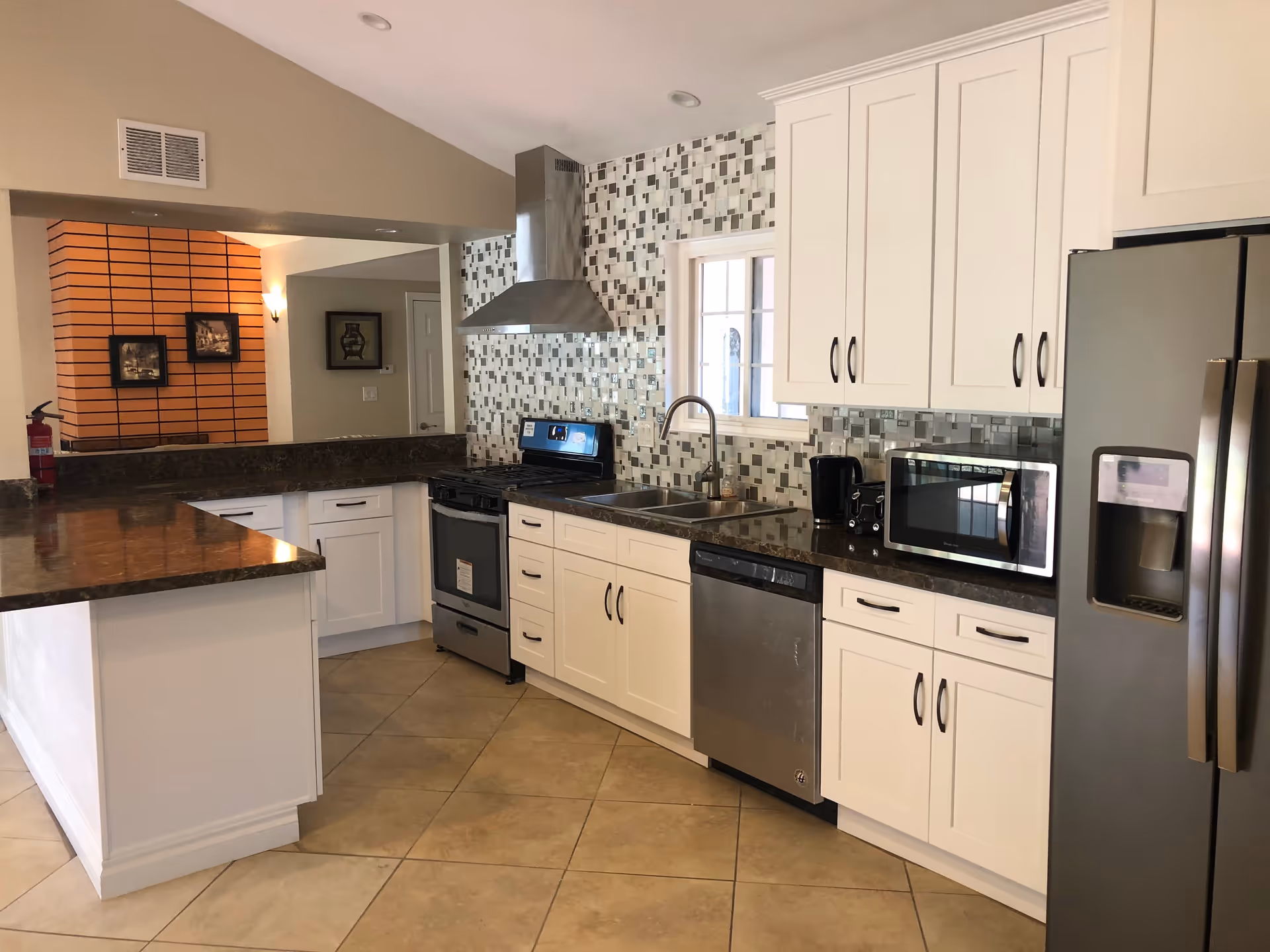 Modern kitchen with white cabinets, stainless steel appliances including a refrigerator, dishwasher, stove, microwave, and a coffee maker. The backsplash features a mosaic tile design in shades of gray and white. There is a window above the sink and a brown granite countertop with a breakfast bar. The floor is tiled in a beige color.
