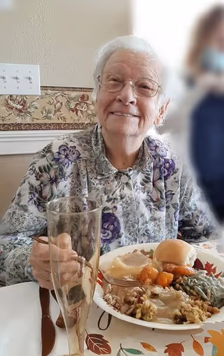 An elderly woman with white hair and glasses is sitting at a table, smiling. She is wearing a floral patterned blouse and is holding a fork. In front of her is a plate of food including mashed potatoes with gravy, green beans, stuffing, a dinner roll, and sweet potatoes. There is also an empty tall glass on the table.