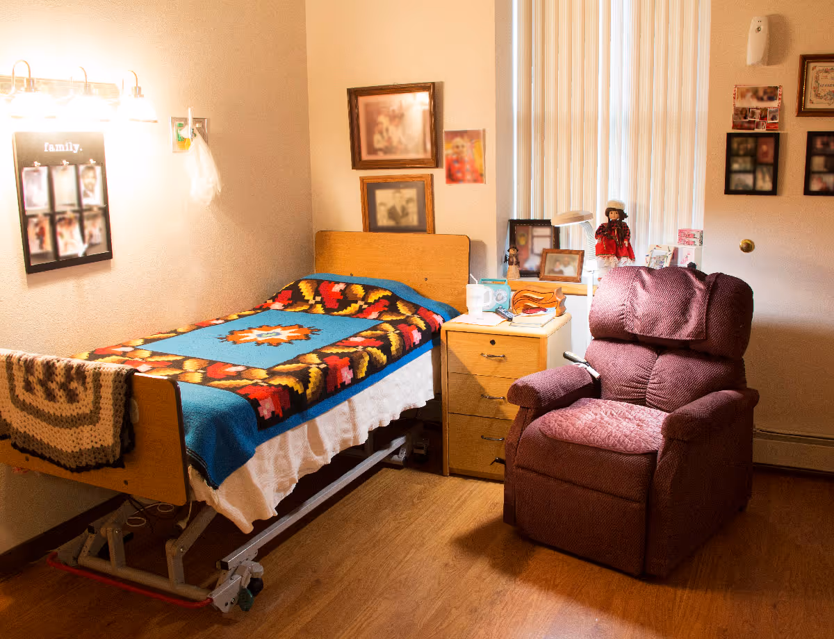A cozy senior bedroom with a single bed covered by a colorful quilt, a wooden nightstand with photos and items, and a burgundy recliner.