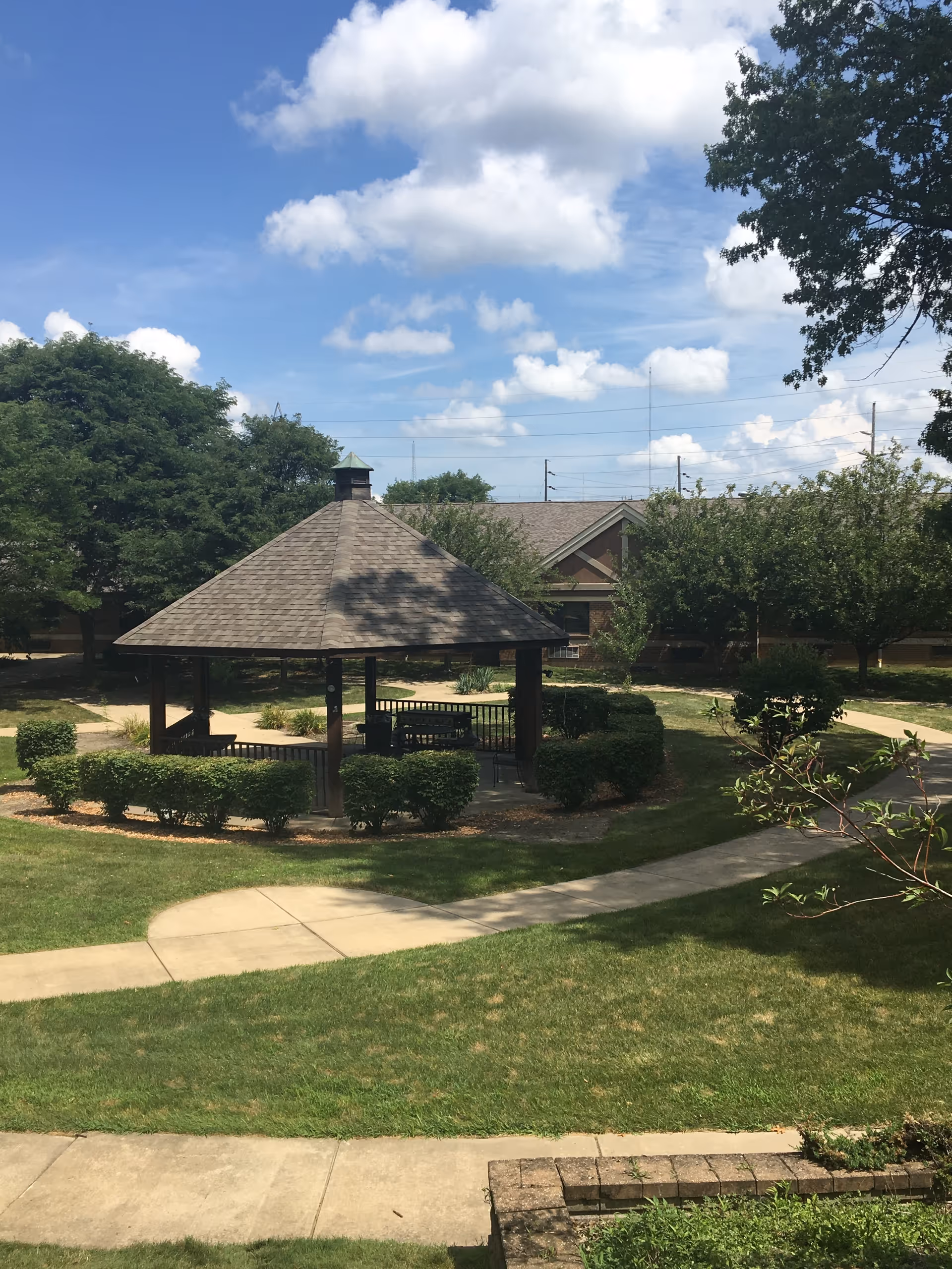 A wooden gazebo sits in a landscaped courtyard with walkways, shrubs, trees, and a building under a blue sky.