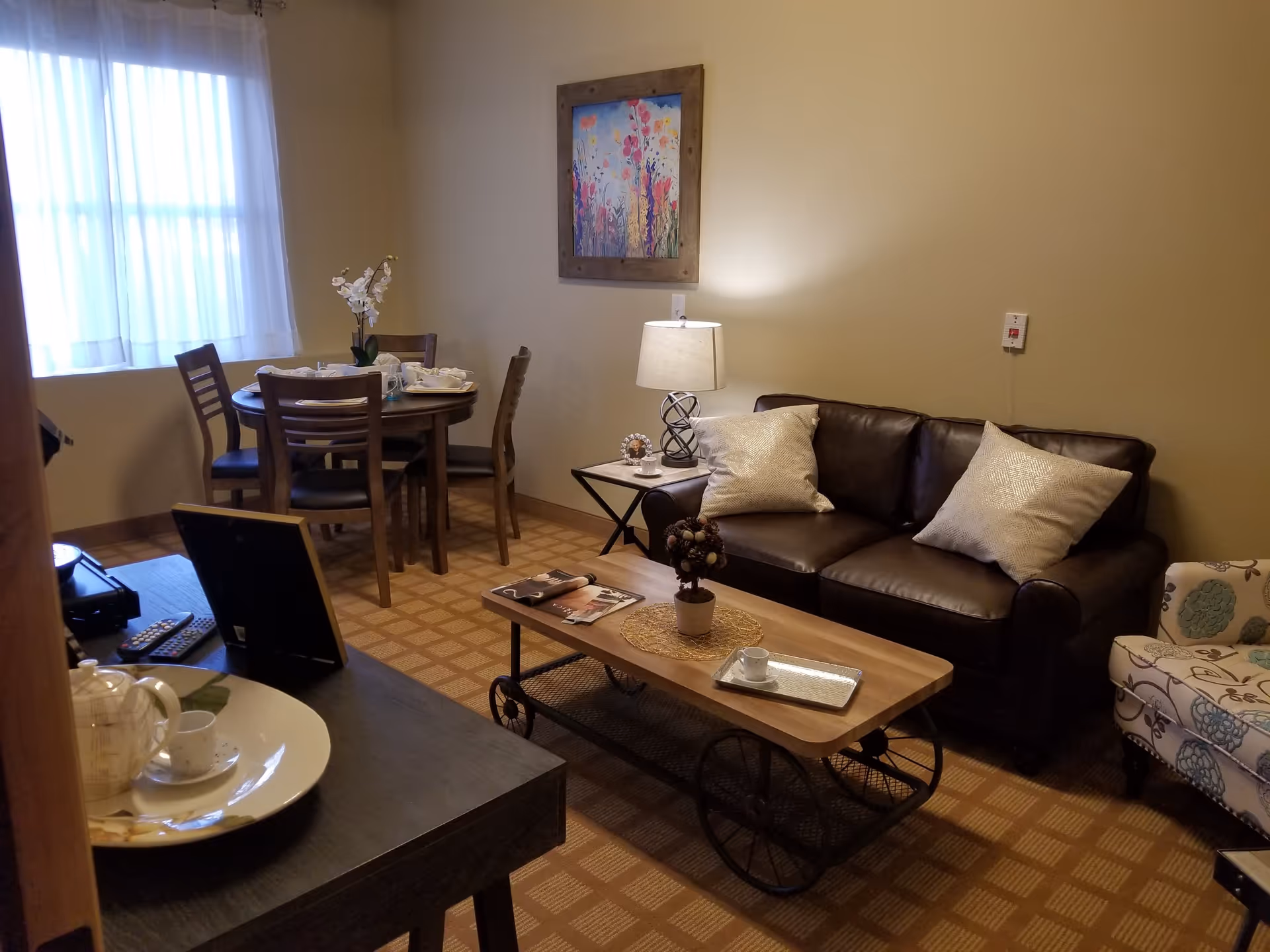Cozy living area with a leather sofa, coffee table on wheels, a small dining table by a curtained window, and decorative accents.