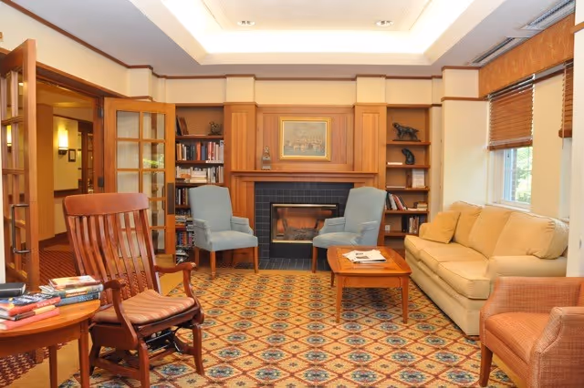 A cozy living room with a patterned carpet, a wooden rocking chair with a cushion, two blue armchairs, a beige sofa, and a wooden coffee table with magazines. There is a fireplace with a wooden mantel and built-in bookshelves on either side. The room has large windows with blinds and warm lighting.