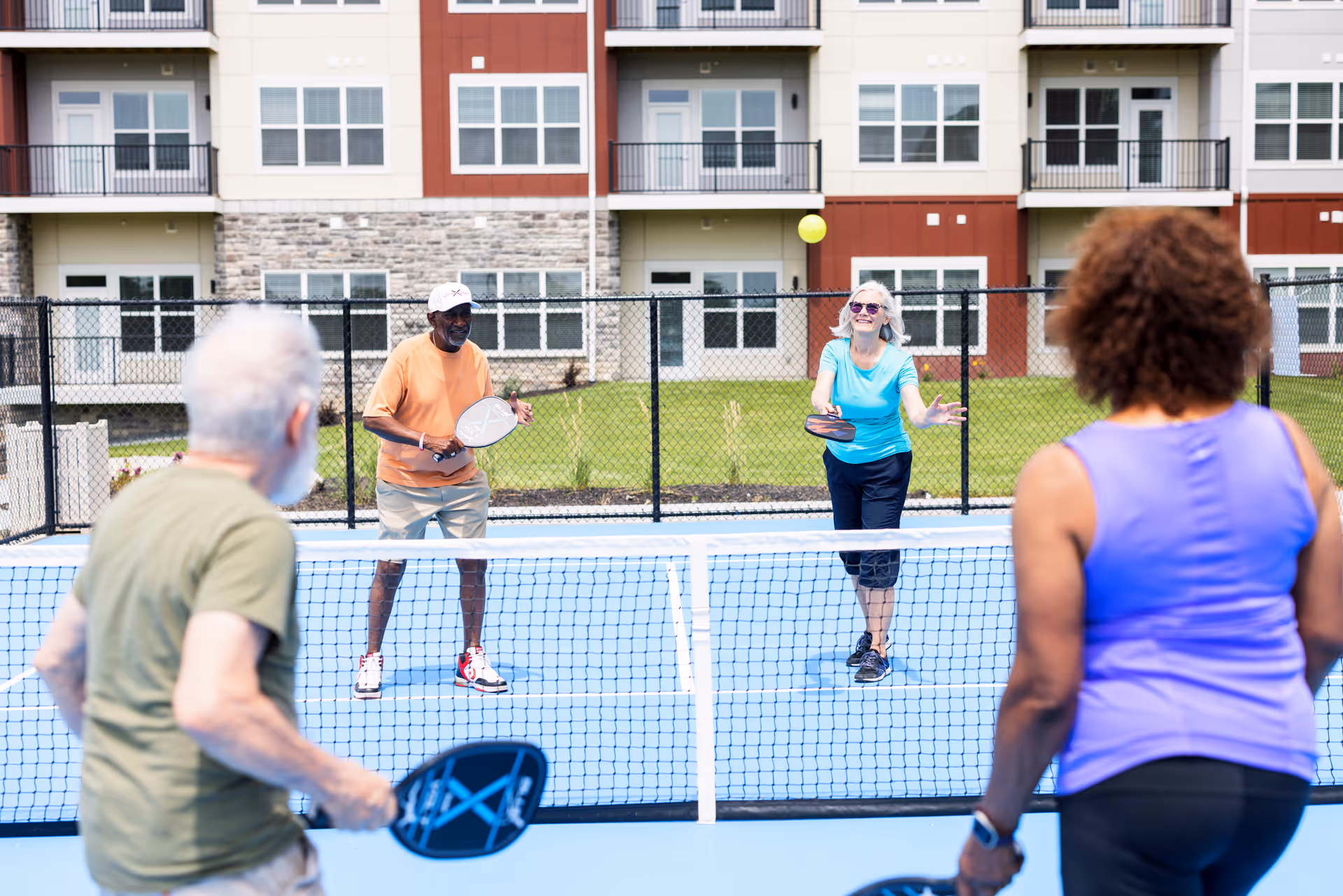 Four seniors playing pickleball on an outdoor court in front of a multi-story residential building. Two men and two women are engaged in the game, holding paddles and hitting a yellow ball over the net.