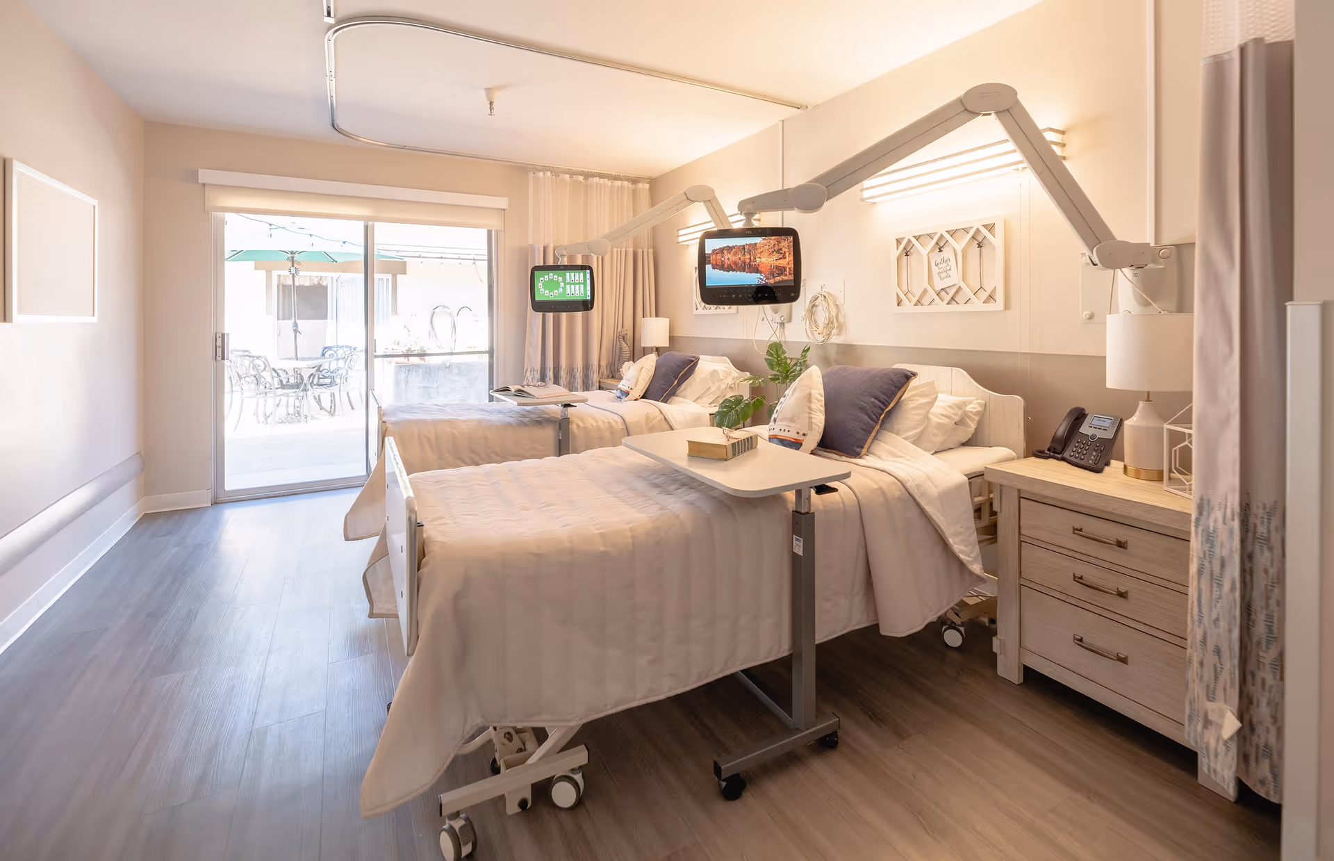 Bright shared resident room with two adjustable hospital-style beds, overbed tables, mounted TVs, nightstands, and a sliding glass door to an outdoor patio.