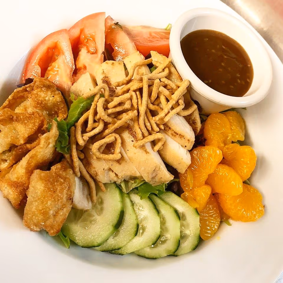 Bowl-style salad with sliced grilled chicken, cucumber slices, mandarin orange segments, tomato wedges, crispy wontons, crunchy noodles and a cup of dressing.