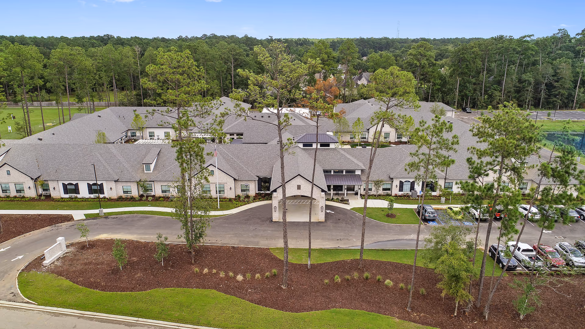 Aerial view of Sage Lake's single-story senior living building with a covered entrance, landscaped grounds, parking lot, and surrounding trees.