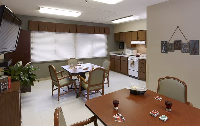 A cozy common area with a small kitchen and dining space. The room features two wooden tables with green cushioned chairs around them. On one table, there are glasses with a dark beverage and a card game laid out. The kitchen area includes wooden cabinets, a microwave, a stove with a range hood, and a paper towel dispenser. A large window with closed blinds and a brown valance lets in natural light. A wall decoration spells out the word 'laugh'. A flat-screen TV is mounted on the left wall above a cabinet with a plant on top.