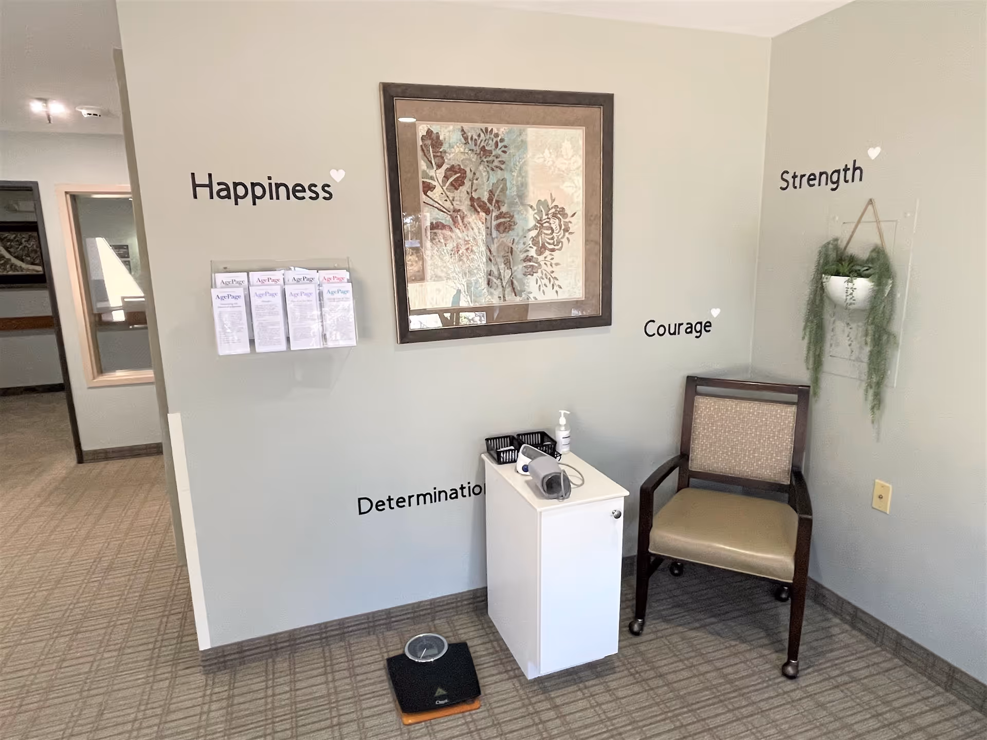 Seating nook in a senior living facility with a chair, small cabinet holding medical items, a floor scale, framed artwork and wall text reading 'Happiness', 'Determination', 'Courage' and 'Strength'.