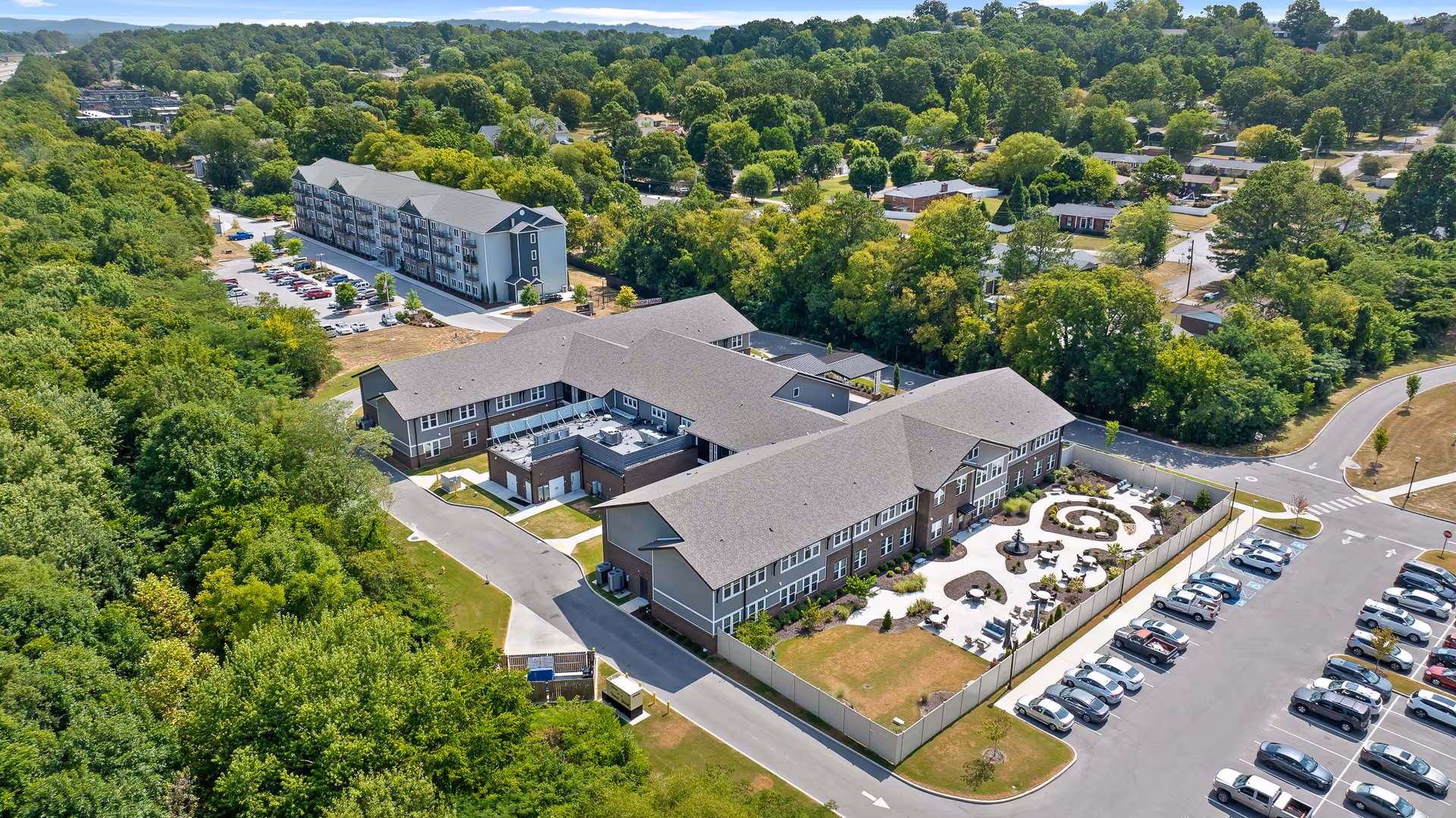 Aerial view of Rockbridge Oaks senior living facility showing multiple connected buildings with gray roofs, a landscaped courtyard with seating and pathways, surrounding greenery, parking lots with cars, and nearby residential neighborhood.