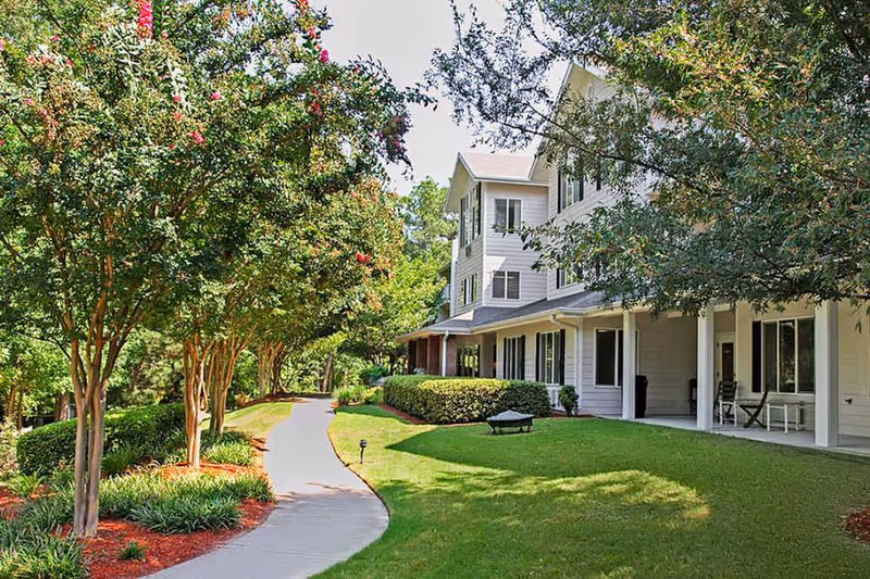 Curved sidewalk through landscaped grounds leading to a multi-story residential building with a covered porch and trees.