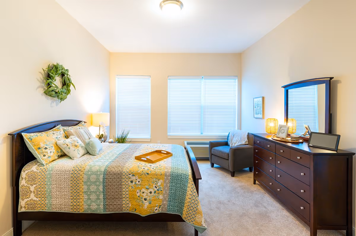 A bright and cozy bedroom with a large bed covered in a colorful quilt with floral and geometric patterns. There are multiple pillows on the bed and a wooden tray placed on top. A green wreath hangs on the wall above the bed. Next to the bed is a nightstand with a lamp and decorative items. Across from the bed is a dark wooden dresser with a large mirror, two lamps, framed photos, and a tablet. A comfortable armchair with a throw blanket is positioned near the windows with white blinds, and a heating/cooling unit is beneath the windows.