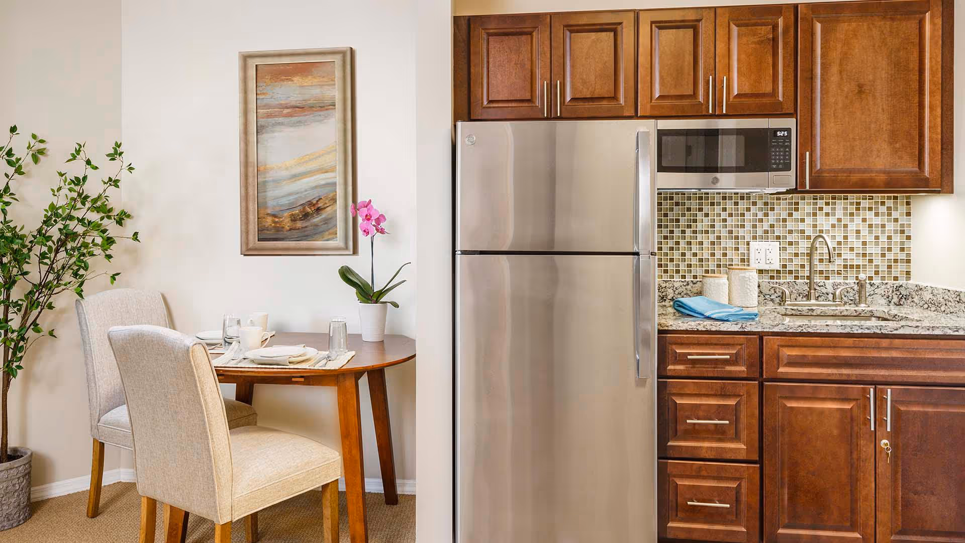 Compact kitchen with stainless steel refrigerator, wooden cabinets, a sink and countertop, and a small round dining table with two upholstered chairs and a potted plant.