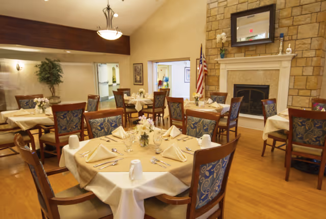 A dining room in Oak Creek Place Assisted Living Community with several round tables covered with beige tablecloths and set with folded napkins, glasses, and silverware. The room features wooden chairs with patterned blue upholstery, a stone fireplace with a mirror above it, an American flag, and warm lighting.