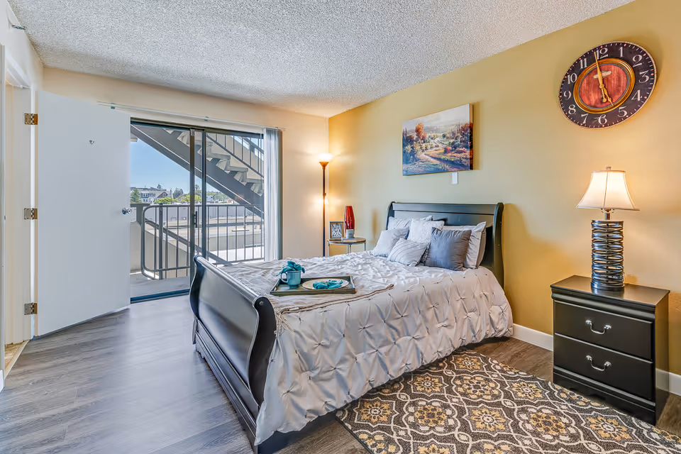 A bright bedroom with a large bed covered in a white comforter and multiple pillows. There is a black nightstand with a lamp on the right side of the bed and a floor lamp in the corner. A colorful painting hangs above the bed, and a large round clock is mounted on the wall. The room has wood flooring with a patterned area rug near the bed. A sliding glass door opens to an outdoor balcony with a metal staircase visible outside.